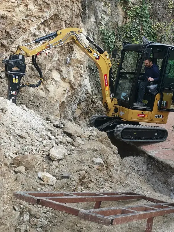 Minibáger CAT s hydraulickým kladivom pri vysekávaní výkopu do skaly